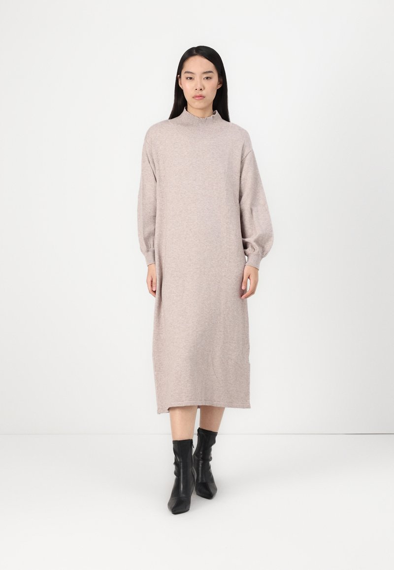 Jasnobeżowa dzianinowa sukienka z wysokim kołnierzem, długimi rękawami i prostym krojem, zestawiona z czarnymi botkami za kostkę. Gładka faktura.