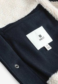 Navy fleece jacket interior with cream sherpa lining, featuring a white label with black text and a metal snap closure.