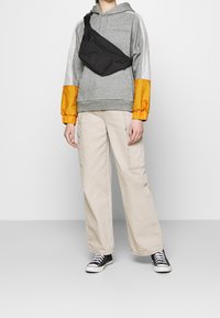 Sweat à capuche gris avec des manches orange, pantalon cargo beige et sac bandoulière noir. Des baskets noires complètent la tenue.