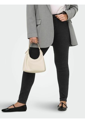 Kvinde i sorte skinny jeans, grå blazer med fine striber, sorte flade sko, der holder en beige lædertaske i håndtaget.