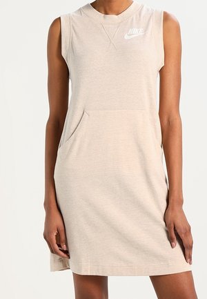 Robe décontractée beige sans manches avec poche avant et logo Nike blanc sur la poitrine, portée par une personne debout sur un fond uni.