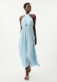 Himmelblaues Maxikleid mit Halterneck, Ausschnittdetail und geraffter Taille. Vertikales Streifenmuster, fließender Stoff und knöchellang. Kombiniert mit Sandalen.
