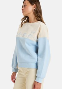 Sweat-shirt bleu clair et beige avec broderie de vagues et poignets côtelés. Tissu doux, coupe décontractée et encolure ras du cou.