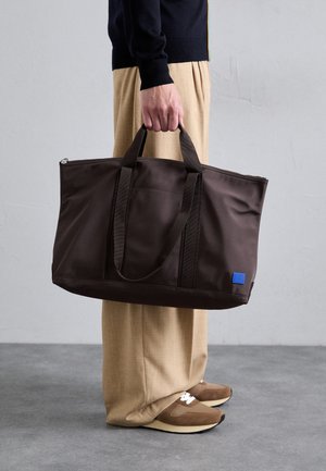 Brown nylon tote bag with dual handles, zip closure, and a blue logo patch. Smooth texture, structured design, spacious interior.