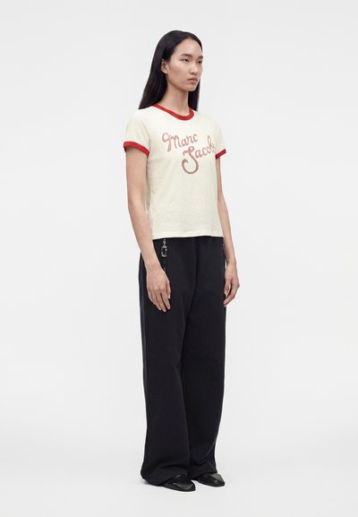 Γυναίκα με κρεμ μπλουζάκι Marc Jacobs με κόκκινο τελείωμα και μαύρο παντελόνι με φαρδύ μπατζάκι στέκεται ελαφρώς στο πλάι μπροστά σε απλό φόντο.