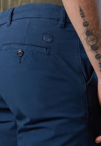 Pantaloni blu navy realizzati in tessuto, con una tasca posteriore con bottone e un dettaglio del logo impresso sopra. Texture liscia e vestibilità su misura.