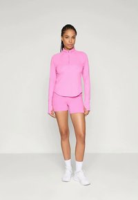 Femme debout portant un haut zippé à manches longues rose vif, un short assorti, des chaussettes blanches et des chaussures de sport blanches sur un fond gris uni.