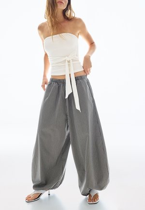 Kobieta ubrana w białą, bezramkową bluzkę z wiązaniem z boku oraz luźne, szerokie spodnie w czarno-białą kratę, do tego białe sandały na obcasie.