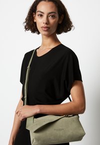 Grüne Wildleder-Crossbody-Tasche mit konischer Form, markenspezifischen Logo-Details und einem schlanken, verstellbaren Trageriemen. Das Modell trägt ein schwarzes Oberteil.