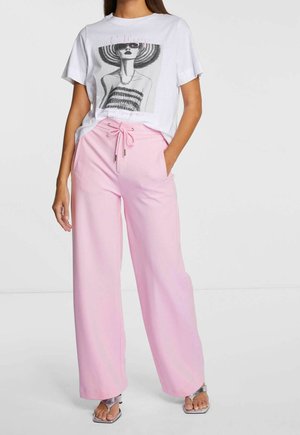Mujer con camiseta gráfica blanca con ilustración de moda, pantalones anchos rosa con cordón y tacones abiertos plateados de pie sobre fondo blanco.