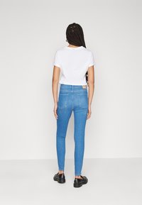 Mujer con trenzas largas, vistiendo una camiseta blanca, jeans ajustados azules y mocasines negros, de pie frente a un fondo liso de color gris claro.