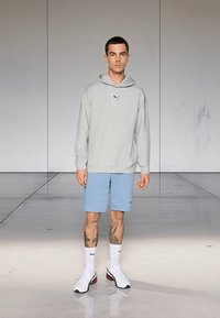 Γκρι φούτερ με κουκούλα και λογότυπο, ανοιχτό μπλε σορτς, λευκές κάλτσες και πολύχρωμα αθλητικά παπούτσια. Το μοντέλο στέκεται μπροστά σε ένα μινιμαλιστικό φόντο.