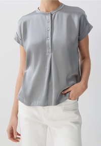 Femme portant un chemisier gris clair à manches courtes avec un plastron à boutons et un pantalon blanc, une main dans la poche, sur un fond uni.