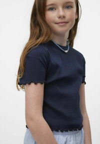 Top corto a righe blu navy con bordi scallop, abbinato a una collana blu e perle, su sfondo bianco semplice.