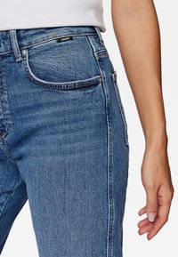 Primer plano de una persona que lleva pantalones de mezclilla azules con el bolsillo delantero visible y una camisa blanca metida por dentro, con el brazo derecho relajado a su lado.