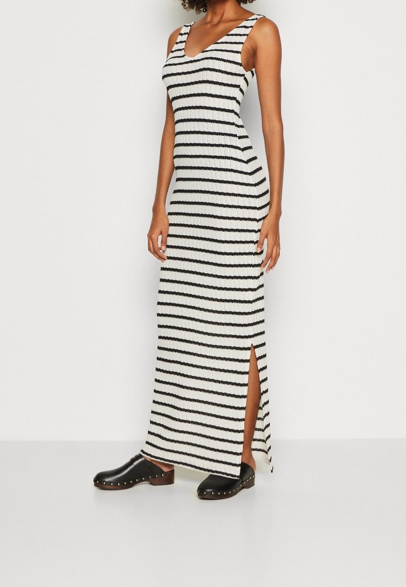 Robe maxi tricot rayée en noir et blanc avec une silhouette moulante, un décolleté arrondi et une fente sur le côté. Associée à des sabots noirs ornés de clous dorés.