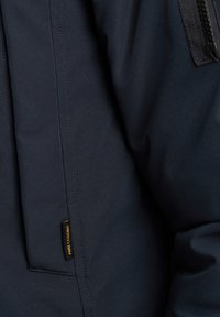 Veste bleu marine foncé avec une texture lisse et mate. Accentuée par une poche zippée et une petite étiquette de logo sur la couture.