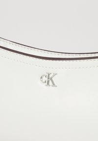 Valkoinen nahkalaukku, jossa on sileä pinta, kontrastivärinen tumman sävyinen reuna ja metallinen "CK"-logo etuosassa.