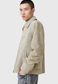 Beige mocka jacka med krage, långa ärmar, dragkedjestängning och sidofickor. Mjuk textur och slät yta med minimal detaljering.