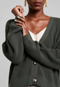 Dunkelgrüne Strickjacke mit tiefem V-Ausschnitt, übergroßen Ärmeln und drei Knöpfen an der Vorderseite. Die strukturierte Stoffoberfläche verstärkt die Tiefe.