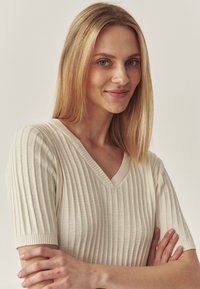 Top de punto acanalado en beige claro con escote en V y mangas cortas. La textura es suave y la tela parece transpirable y cómoda.