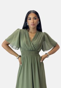 Robe vert olive avec des manches courtes évasées, un décolleté en V et une taille froncée. Le tissu est léger et fluide.