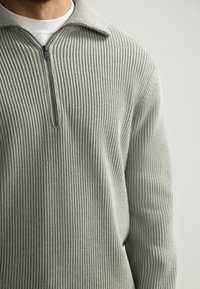 Man in een lichtgrijze geribde zip-up sweater over een witte t-shirt, weergegeven van hals tot midden torso tegen een effen achtergrond.