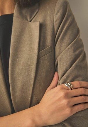 Person wearing a beige blazer with arms crossed, showing two thick metallic rings on their right hand's index finger.