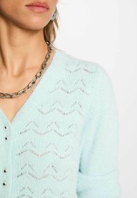 Cardigan lavorato a maglia azzurro chiaro con una texture morbida, scollo a V e motivo a onde con dettagli in bottone metallico. Collana in argento in vista.