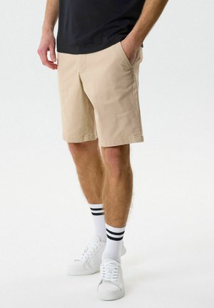 Egy férfi bézs térd alatti nadrágot, fehér tornacipőt és fekete csíkos fehér zoknit visel, egyik kezét zsebbe dugva áll.