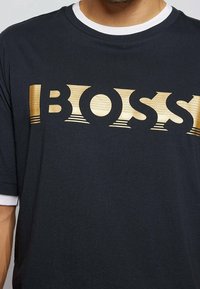 Mörkblå t-shirt med korta ärmar, som har en framträdande guld "BOSS"-logotyp i ett horisontellt mönster med texturerade detaljer.