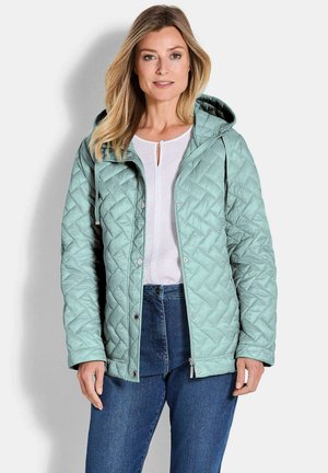Lichtgroene gewatteerde jas met een capuchon, met drukknopen en een ritszak, gedragen over een wit shirt en een spijkerbroek. Gladde textuur.