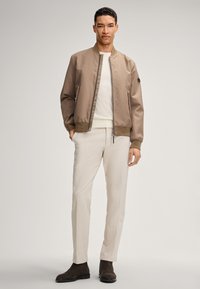 Giacca bomber color sabbia con finitura liscia, chiusura con zip e polsini a coste, abbinata a pantaloni in lino beige chiaro e stivali in suede marrone scuro.