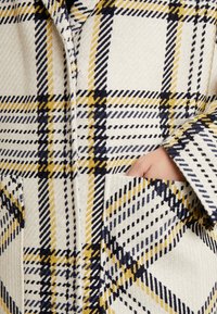 Main dans la poche d'une veste crème avec un motif à carreaux jaune, noir et bleu marine, montrant un tissu texturé et des détails de poche cousus.