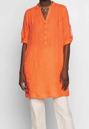 Tunic in lino arancione con scollo a V, maniche arrotolabili e cinque bottoni sul colletto. Abbinata a pantaloni color crema.