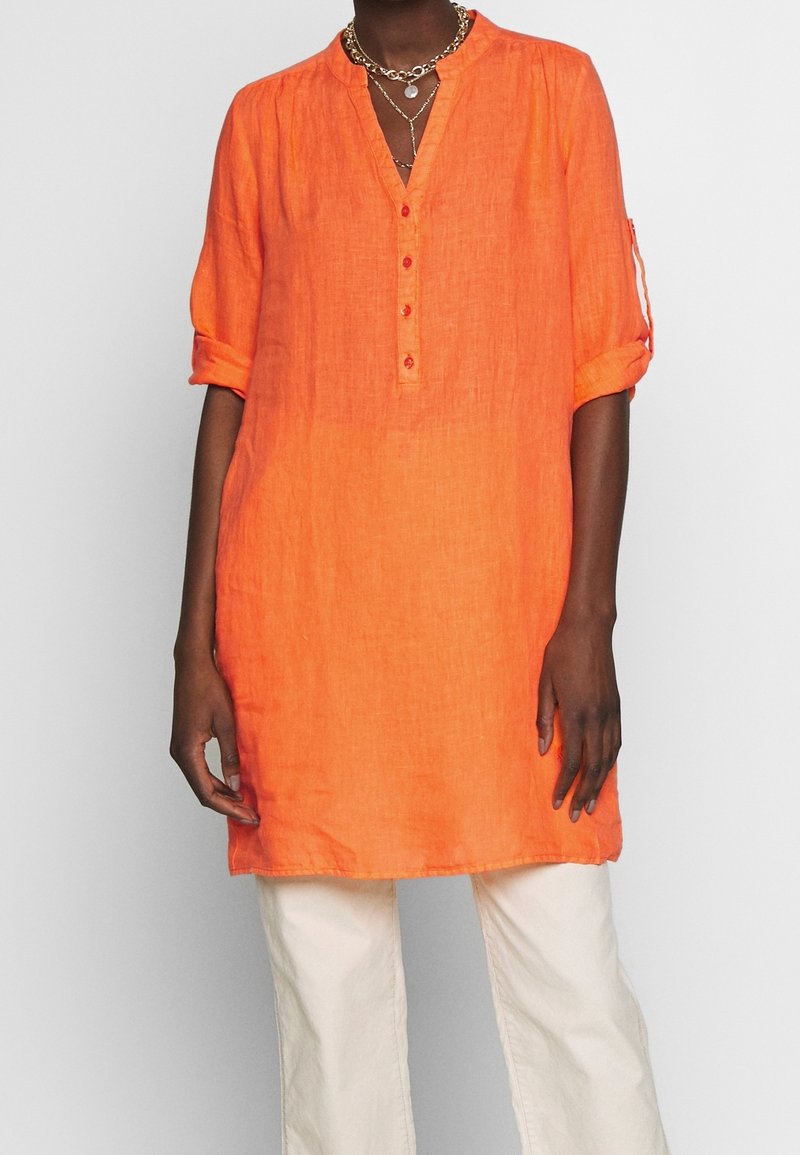 Tunic in lino arancione con scollo a V, maniche arrotolabili e cinque bottoni sul colletto. Abbinata a pantaloni color crema.