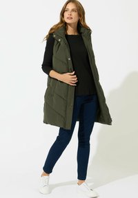 Olivgrüne gepolsterte Weste mit hohem Kragen, ausgestattet mit einem Reißverschluss und Druckknöpfen. Getragen über einem schwarzen Oberteil und dunklen Jeans mit weißen Sneakern.