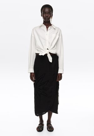 Mujer de piel oscura con camisa blanca anudada, falda midi negra con diseño floral texturizado y zapatos negros sin cordones con adornos de cuentas coloridas.