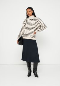 Femme portant une jupe plissée noire, des bottes noires montantes jusqu'aux genoux et un pull duveteux à rayures blanches et noires, tenant une pochette noire devant un fond blanc.