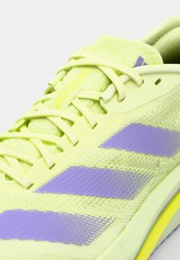 Scarpa sportiva verde chiaro con tomaia testurizzata, strisce di accentuazione viola e marchio giallo. Caratterizzata da lacci sottili e suola ammortizzata.