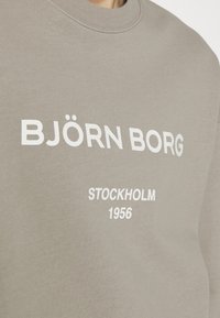 Gros plan sur un sweat-shirt taupe avec le texte blanc "BJÖRN BORG" et un texte plus petit "STOCKHOLM 1956" sur la poitrine.