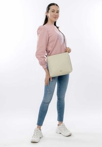 Borsa a tracolla beige chiaro con una texture liscia, caratterizzata da un logo sottile. Il modello indossa un maglione rosa e jeans strappati blu chiaro.