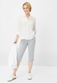 Camicia bianca di lino con bottoni, pantaloni grigi a vita alta, sneakers bianche con suola marrone chiaro. Giacca tenuta nella mano sinistra, capelli pettinati corti.