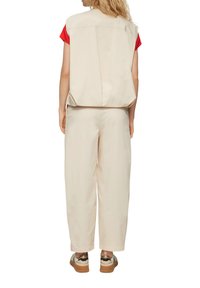 Beige ärmelloses Oberteil und weite Hose aus leichtem Stoff. Roter Akzent auf dem Oberteil, legere braun-goldene Schuhe mit strukturierter Sohle.