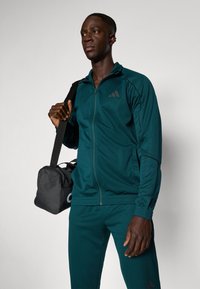 Teal atletski trenirka z zip- up jakno in slim-fit hlačami. Teksturovana tkanina, črne poudarjene črte in logo. Drži črno športno torbo.