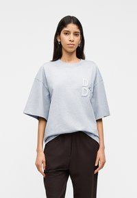 Γυναίκα με μακριά σκουρόχρωμα μαλλιά που φοράει ασημένια σκουλαρίκια κρίκους, φαρδιά ανοιχτογκρι μπλούζα με ανάγλυφα γράμματα και σκουρόχρωμο παντελόνι πάνω σε λευκό φόντο.