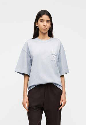 Donna con lunghi capelli scuri che indossa orecchini a cerchio argentati, una maglietta grigia chiara oversize con lettere in rilievo e pantaloni scuri su sfondo bianco.