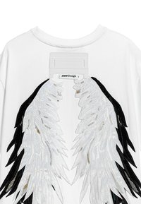 Camiseta blanca con un diseño de alas de ángel bordadas en negro y plata en la parte posterior, que presenta una etiqueta rectangular de MWM Design en el cuello.