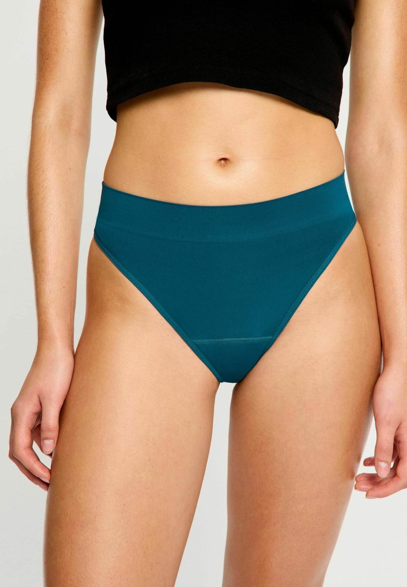 Femme portant des sous-vêtements sans couture taille haute turquoise et un crop top noir, se tenant debout avec les bras détendus le long du corps.