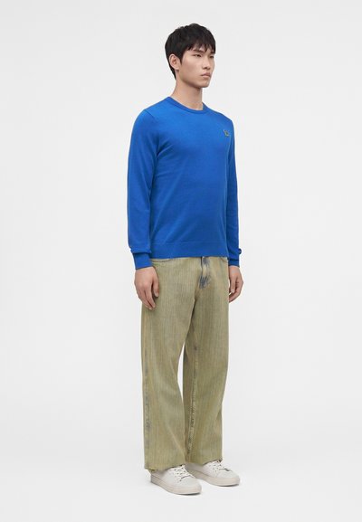 Jeune homme debout portant un pull bleu vif, un pantalon beige ample et des baskets blanches sur un fond clair uni.